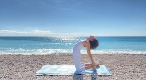 Rosa, la profesora de RYLS YOGA, realiza una postura de camello sobre una esterilla de yoga junto al resplandeciente mar azul, capturando la esencia de la armonía con el entorno natural.